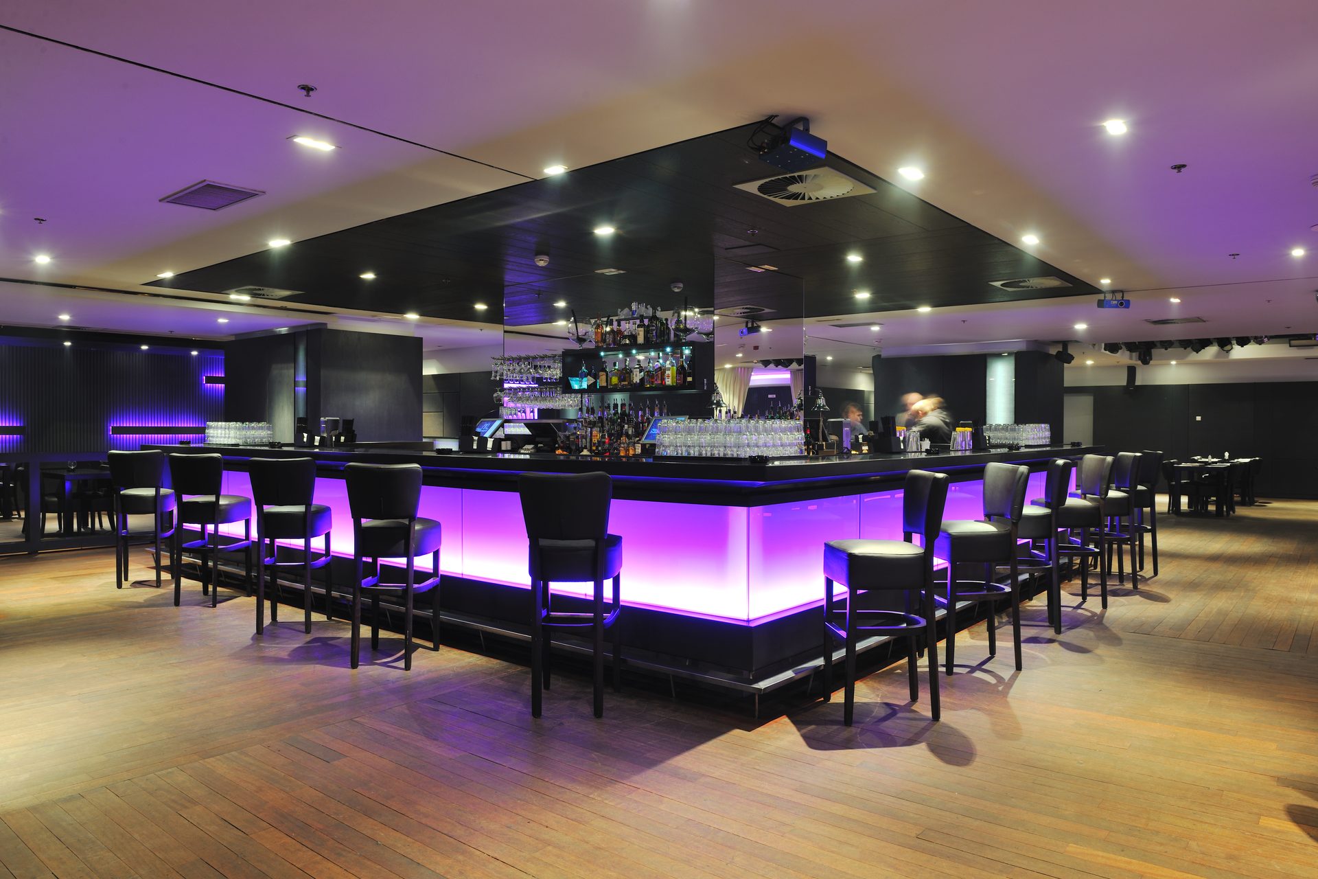 A large bar with its counter front lit purple from within, black leather bar stools around it and downlights set in a dark ceiling panel above