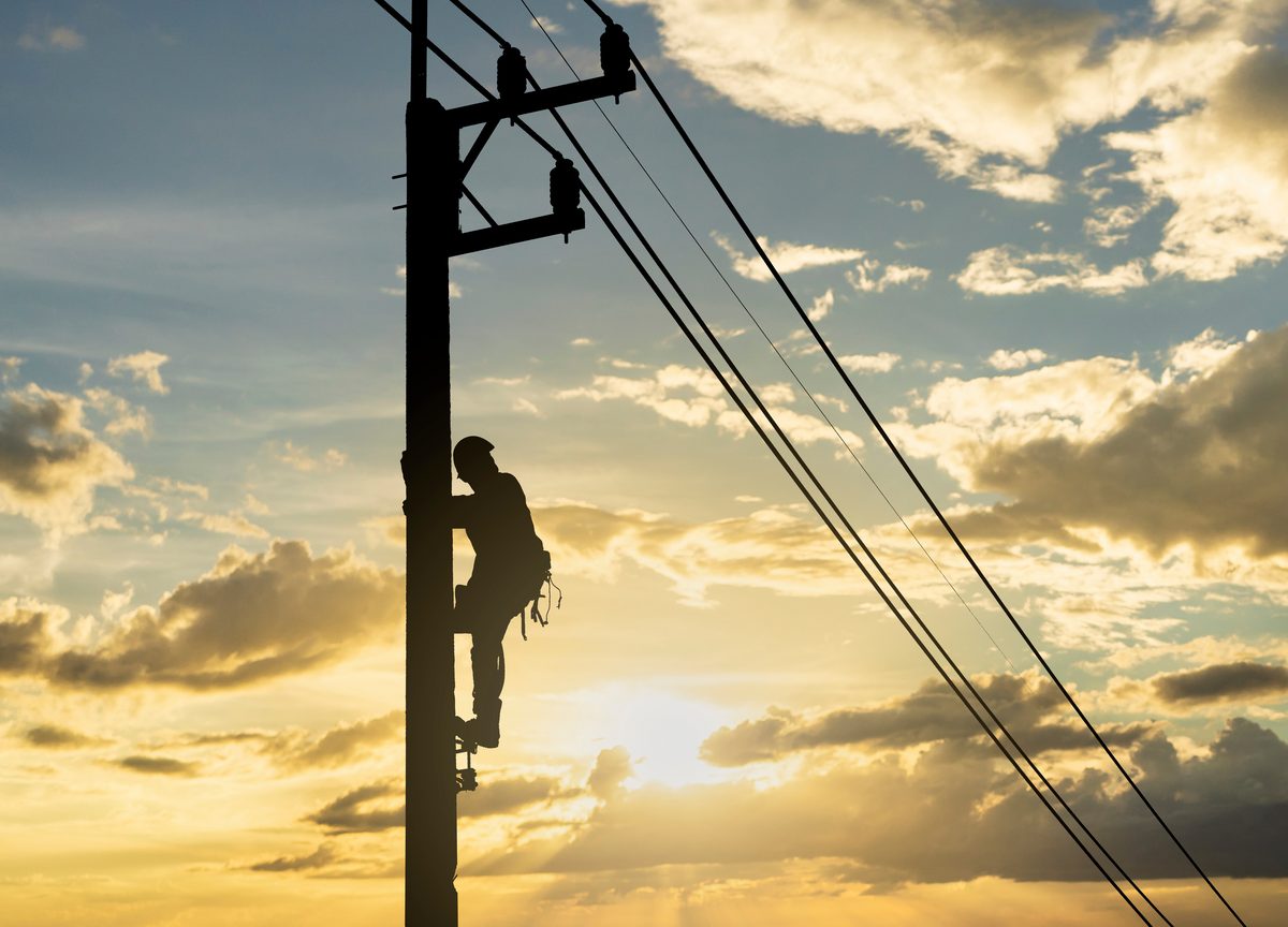 Silhouette of a lineworker climbing a timber power pole carrying overhead lines, against a cloudy golden sunset