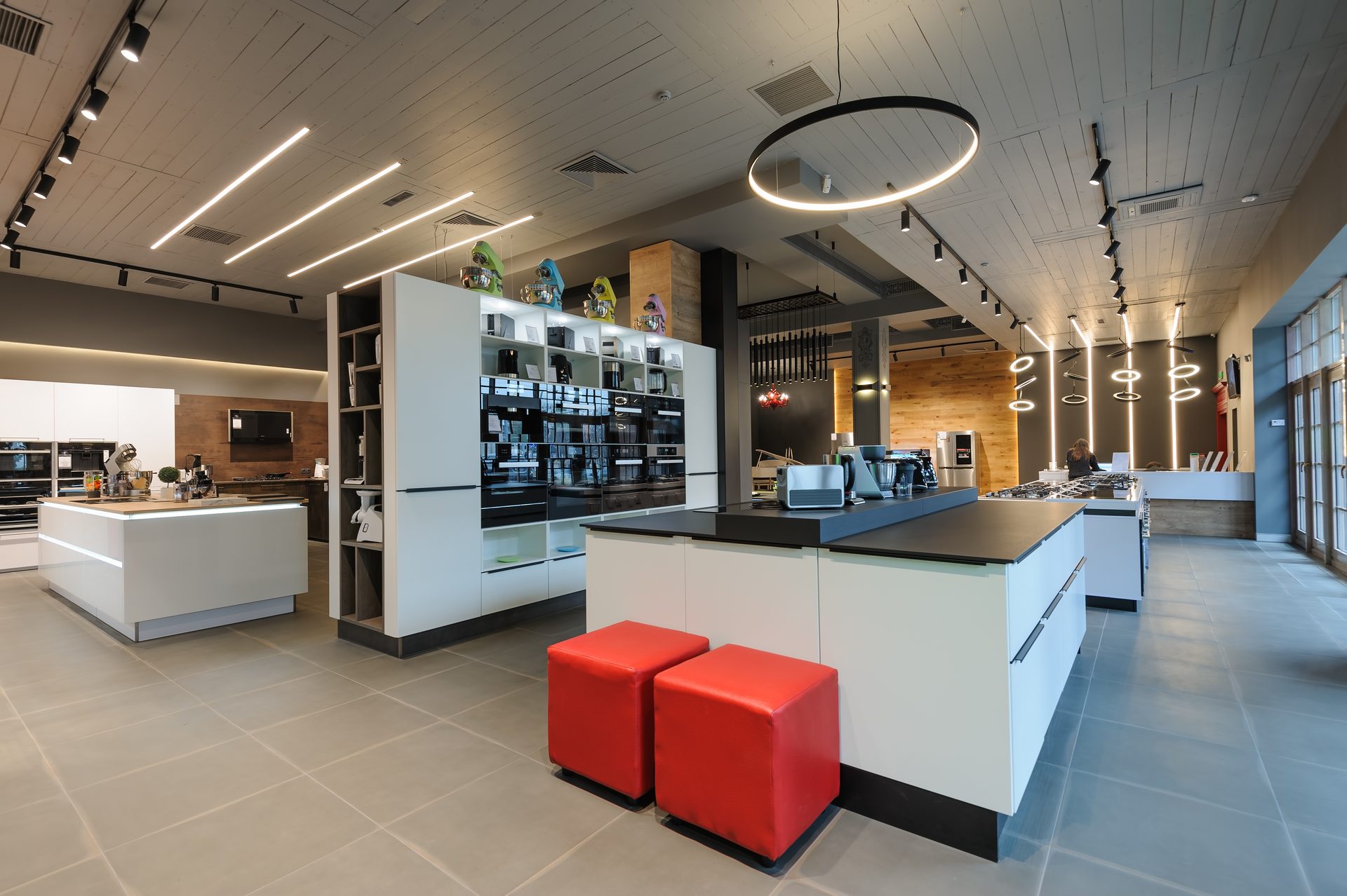 A kitchen appliance showroom lit by black track spotlights, linear LED strips in a timber-board ceiling and a large ring pendant, with two red cube stools beside a display island